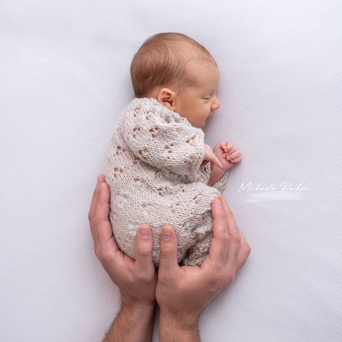 Schlafendes Neugeborenes in hellem Strickoutfit liegt seitlich zusammengekauert, gehalten von zwei schützenden Händen auf weißem Untergrund.